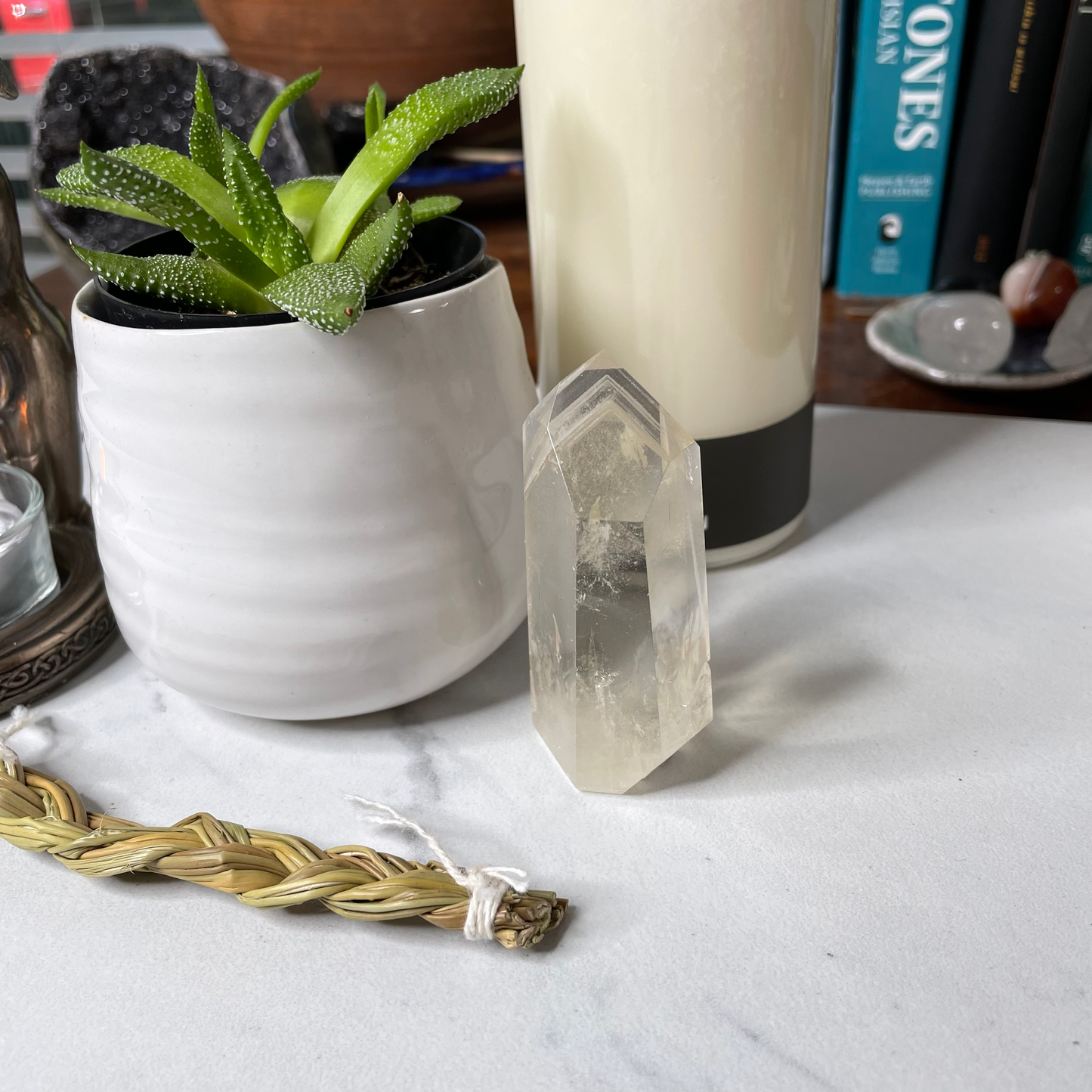 Freya's Haven | Metaphysical & Crystal shop | A pale yellow stone called Citrine has been polished into a standing point. It is displayed with a succulent in a white pot, a goddess tea light candle holder with lit candle and a braid of sweet grass on a white marble tile. there are other crystals, a plant and books in the background.