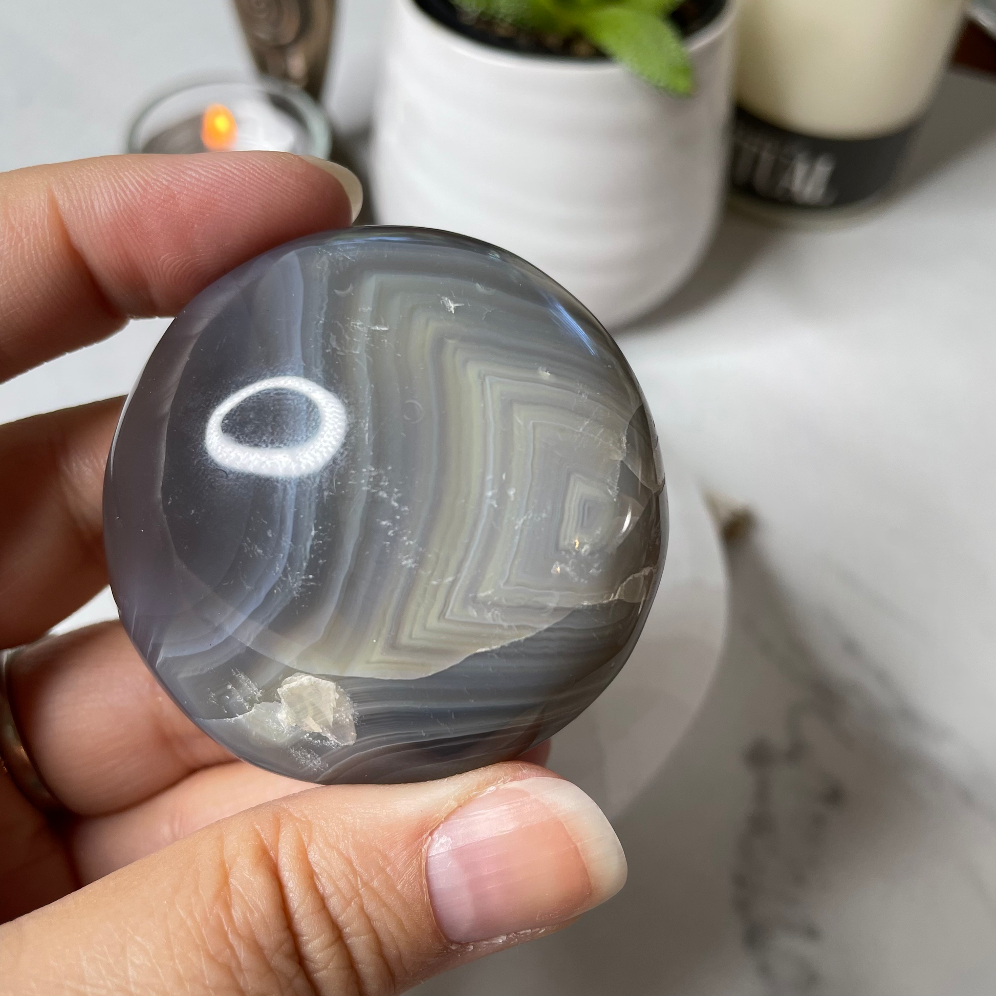 Freya's Haven | Metaphysical & Crystal shop | Three blue and white banded palm stones of Orca Agate are stacked in a white Selenite bowl on a white marble tile. There are candles and a pot in the background.
