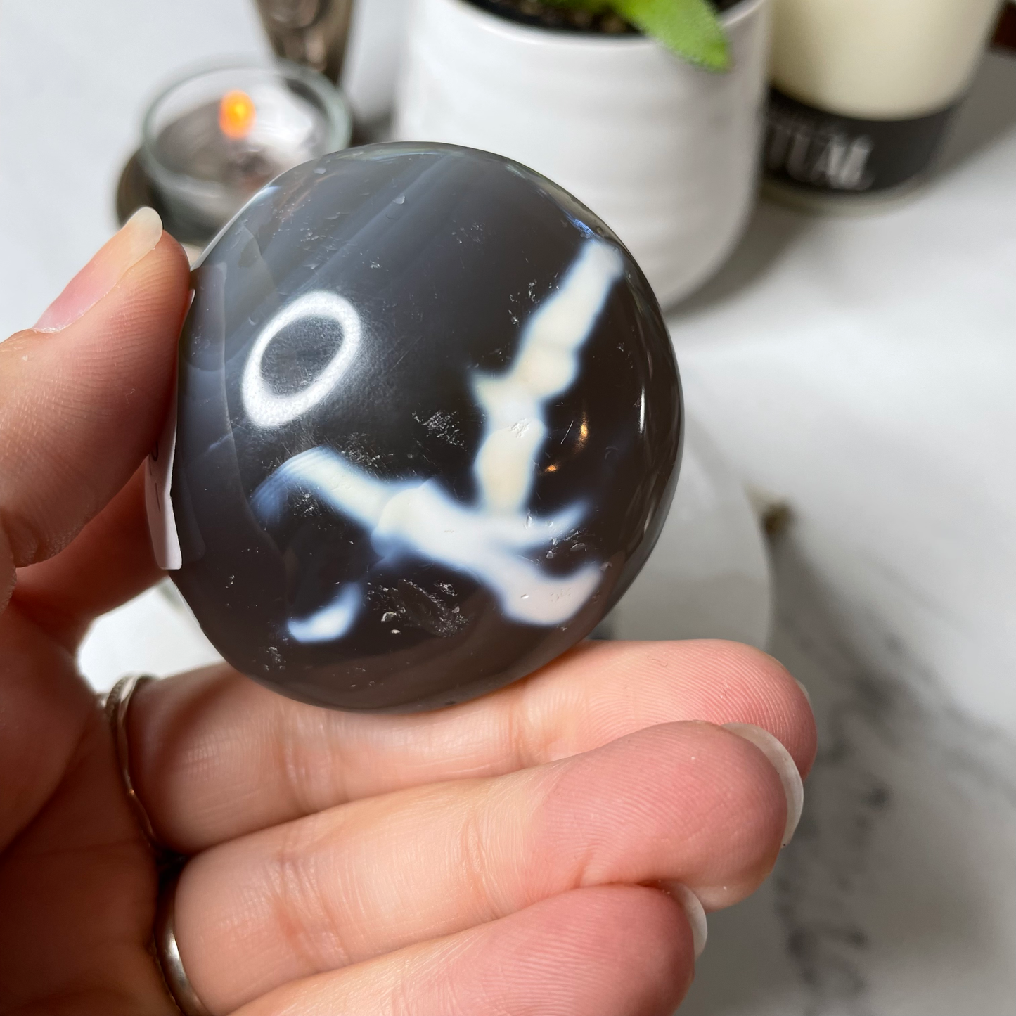 Freya's Haven | Metaphysical & Crystal shop | Three blue and white banded palm stones of Orca Agate are stacked in a white Selenite bowl on a white marble tile. There are candles and a pot in the background.