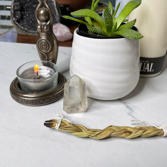 Freya's Haven | Metaphysical & Crystal shop | A pale yellow stone called Citrine has been polished into a standing point. It is displayed with a succulent in a white pot, a goddess tea light candle holder with lit candle and a braid of sweet grass on a white marble tile. there are other crystals, a plant and books in the background.